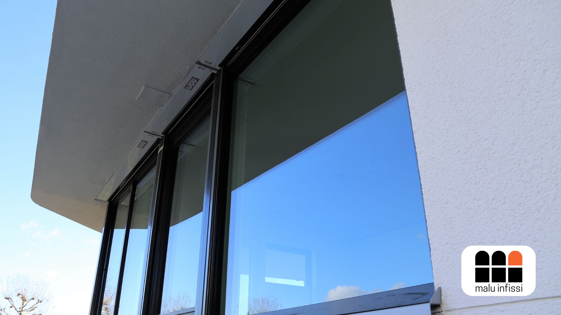 Fotografia in formato orizzontale che mostra una facciata esterna di un edificio moderno. In primo piano, si vedono grandi vetrate a tutta altezza con infissi in alluminio nero e vetri riflettenti che specchiano il cielo azzurro. La parete esterna è bianca e presenta una texture leggermente ruvida. In basso a destra è visibile il logo di Malu Infissi. L’angolo di ripresa dal basso verso l’alto enfatizza la pulizia delle linee architettoniche e l’ampiezza delle superfici vetrate.